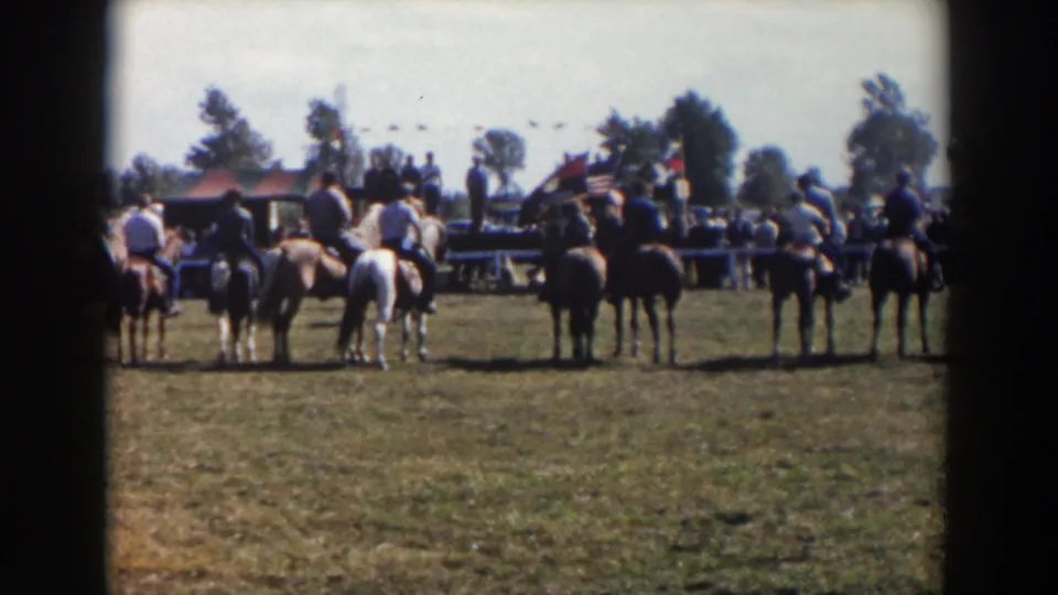 The home-movie frame shows riders and horses from behind in a grassy Wheeling, Ohio setting, with spectators and flags beyond them.