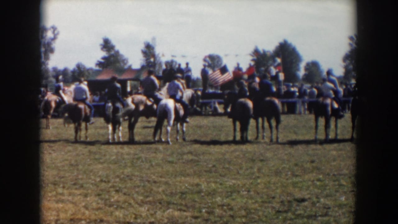 1957 Wheeling, Ohio home-movie frame showing mounted men on brown and white horses near spectators and flags in a grassy event area.