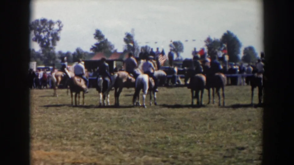 Mounted riders remain grouped in the grassy Wheeling, Ohio event scene, with flags, spectators, trees, and a low building behind them.