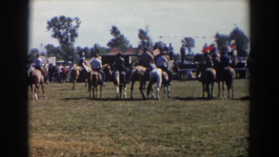 The 1957 home-movie view shows riders and horses in a grassy field, with spectators clustered near flags and trees in the distance.