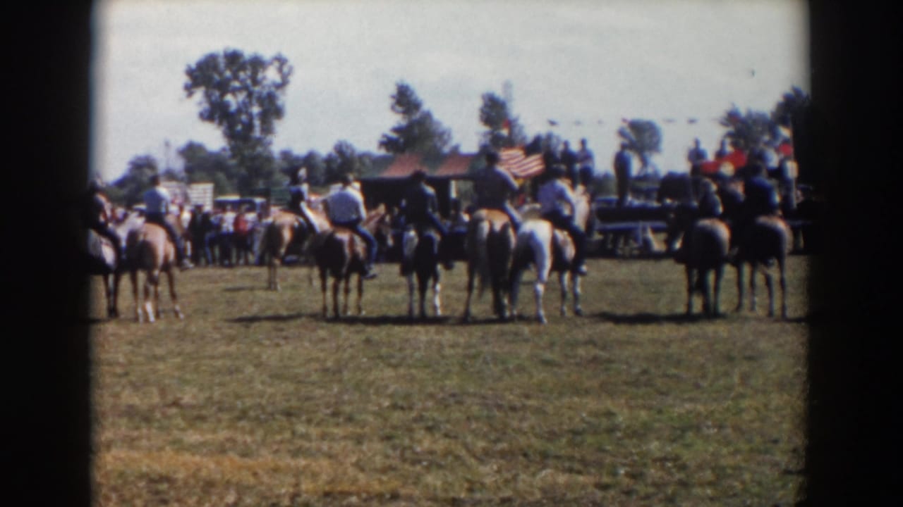 1957 Wheeling, Ohio home-movie frame showing riders on horseback gathered on grass with spectators, trees, and an American flag beyond them.