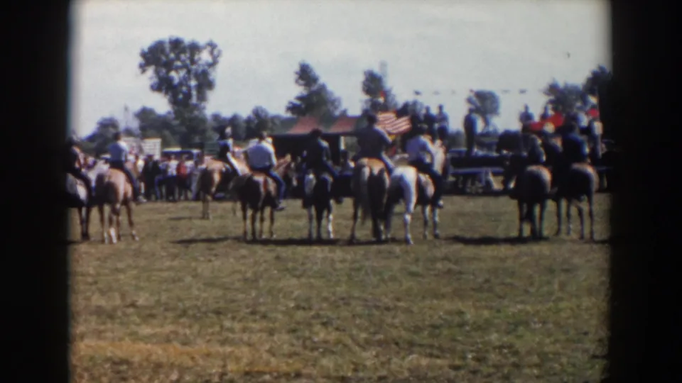 1957 Wheeling, Ohio home-movie frame showing riders on horseback gathered on grass with spectators, trees, and an American flag beyond them.