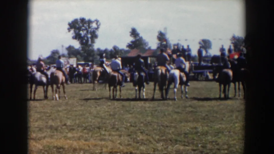 Mounted riders remain lined up on a grassy field at the Wheeling, Ohio event, with distant spectators, trees, and an American flag visible.