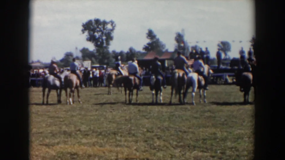 The home-movie view holds on riders and horses from behind as they wait near spectators at an outdoor 1957 Wheeling, Ohio horse event.