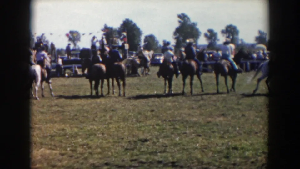 A wider 8mm frame shows horseback riders spaced across the grassy field, with spectators, vehicles, trees, and event activity in the distance.