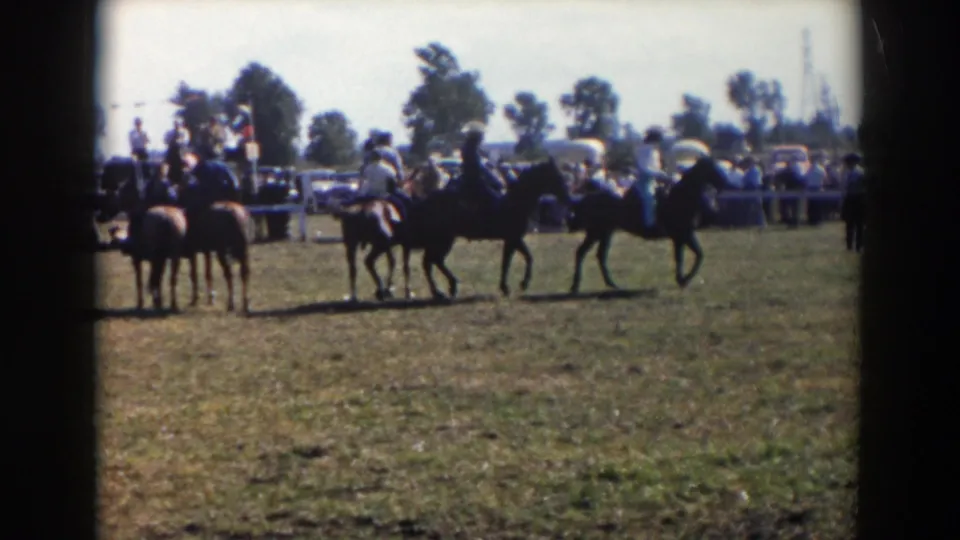 Several mounted riders start trotting across a grassy field during the Wheeling, Ohio event, with spectators, cars, and trees in the background.
