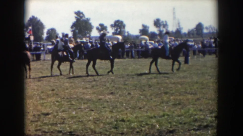 Two mounted riders move through the frame in the 1957 outdoor Ohio scene, with a fenced audience and parked mid-century cars behind them.