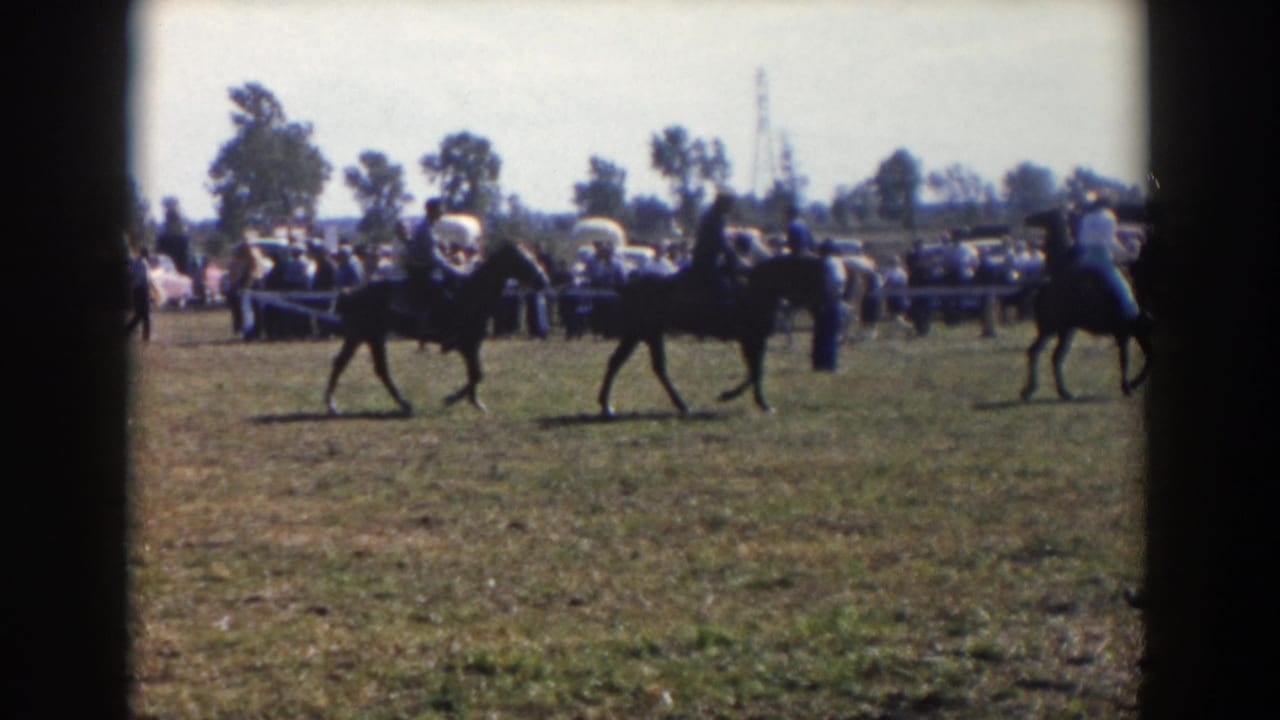 1957 Wheeling, Ohio home-movie frame showing riders on dark horses in an outdoor horse show with spectators and cars behind them.