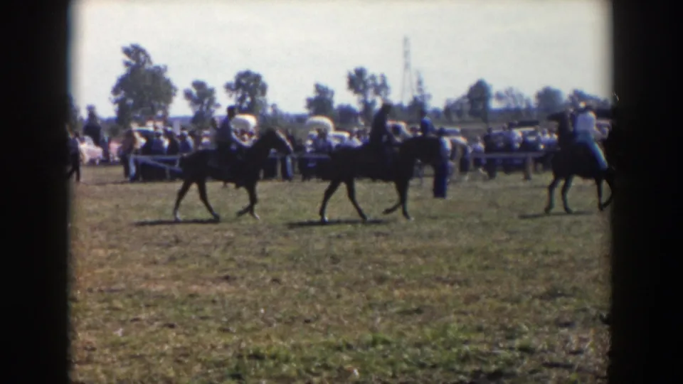 1957 Wheeling, Ohio home-movie frame showing riders on dark horses in an outdoor horse show with spectators and cars behind them.