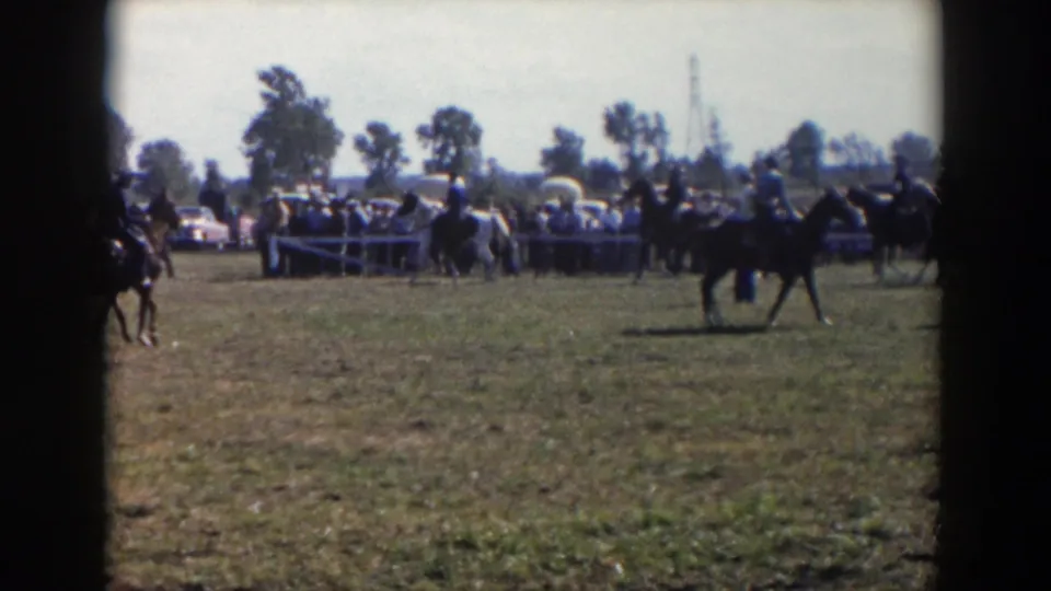 A rider on a dark horse crosses the show field as distant spectators and mid-century cars line the background.