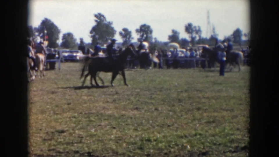 1957 Wheeling, Ohio home-movie frame showing riders on horses in a grassy event field with spectators and cars in the background.