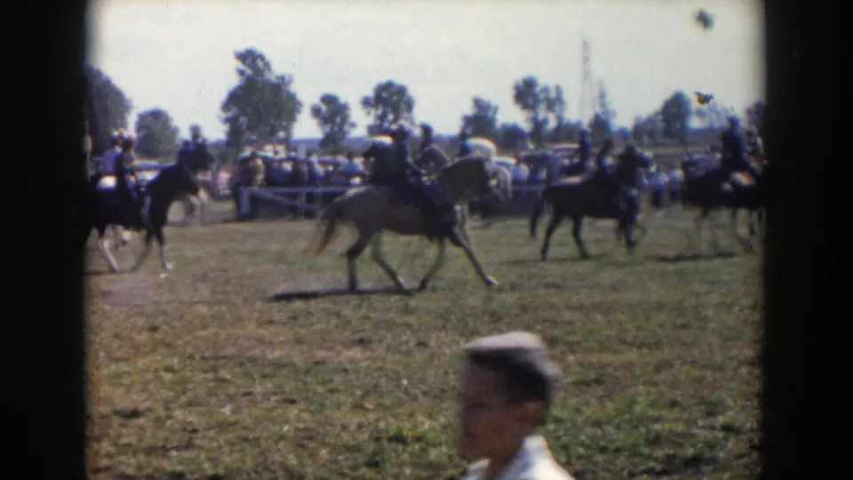 A home-movie view of riders on horses crossing the field, with spectators, cars, and a foreground passerby visible.