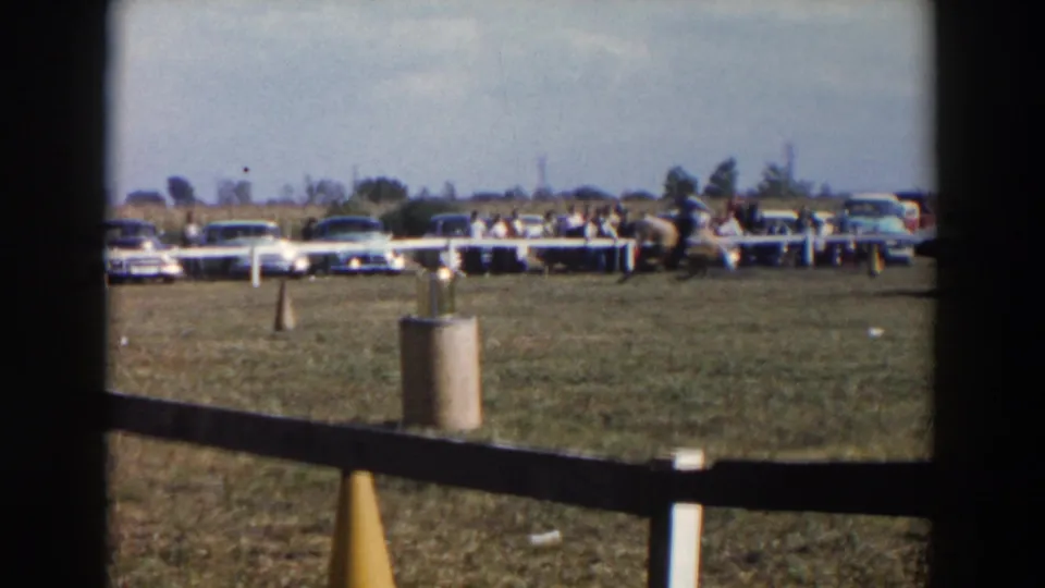 The field edge shows spectators and parked cars behind a simple barrier, with horses and riders farther across the grounds.