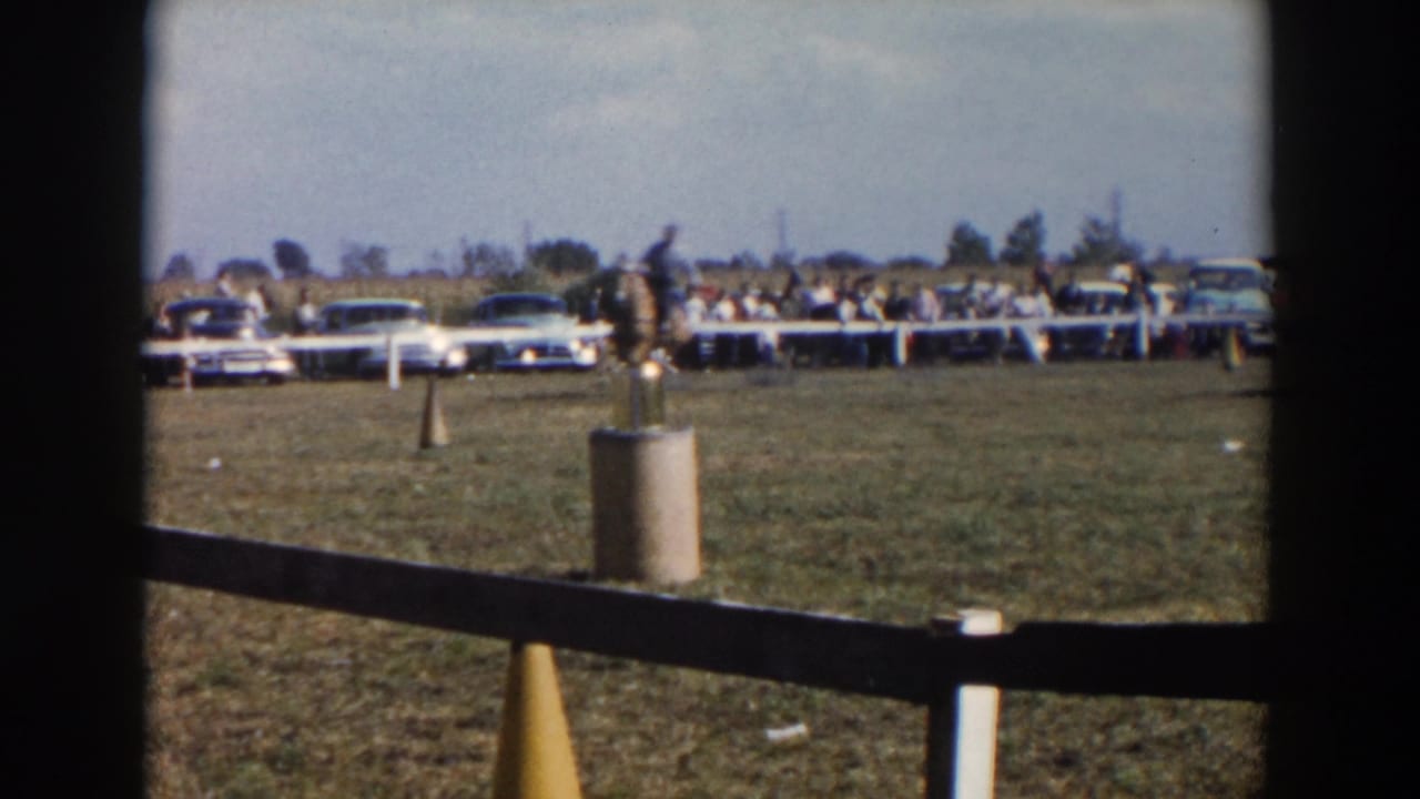 Distant rider on horseback crossing a grassy field with spectators and parked cars beyond a white fence.