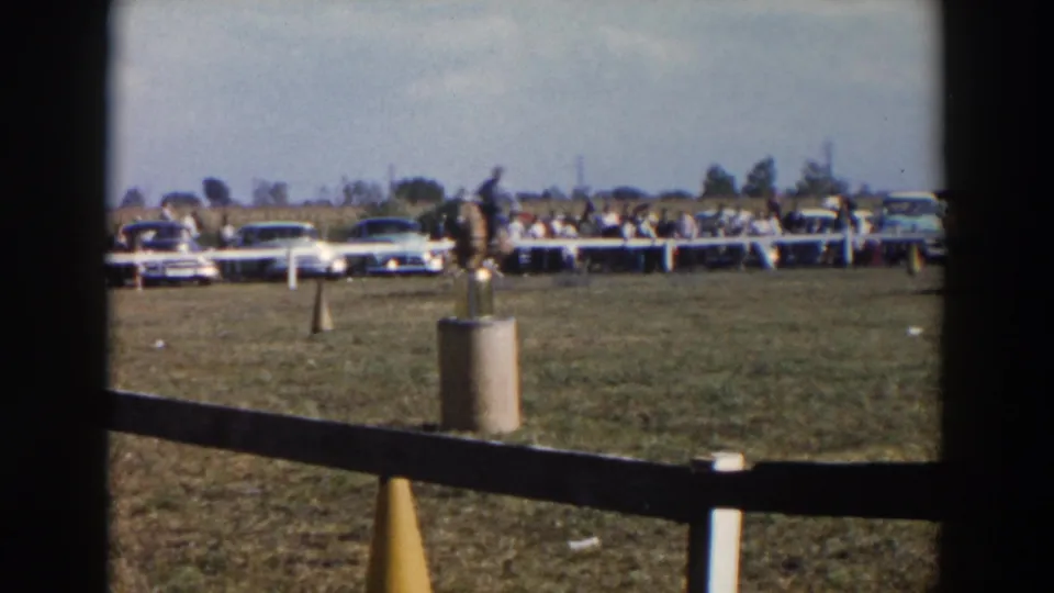 1957 Wheeling, Ohio home-movie frame showing a distant horse and rider on a grassy practice field with spectators and classic cars in the background.