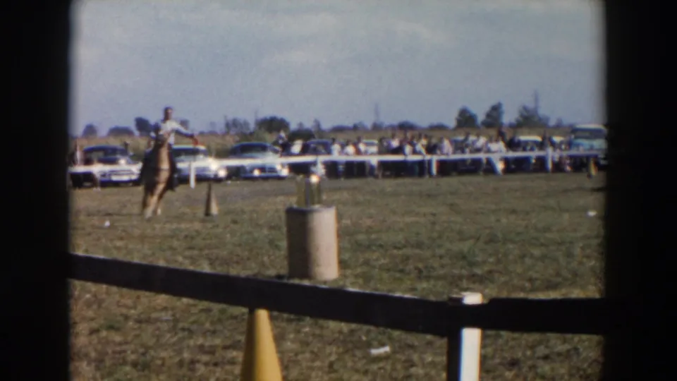 The rider guides the horse toward a cylindrical marker during a 1957 Wheeling, Ohio equestrian practice scene, with spectators and parked cars behind the rail.