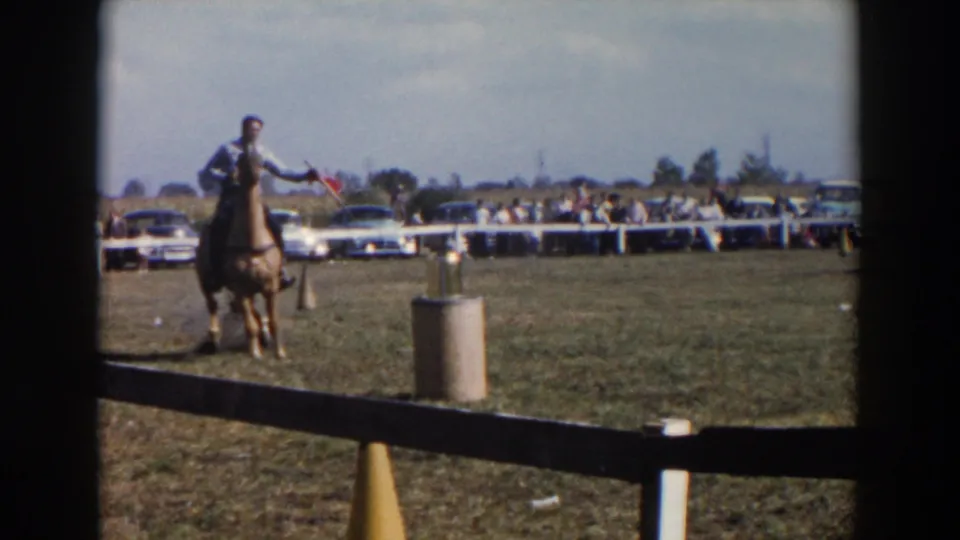 A light-shirted rider on horseback nears a marker on the grassy Wheeling practice field, with mid-century cars and spectators visible beyond the fence.
