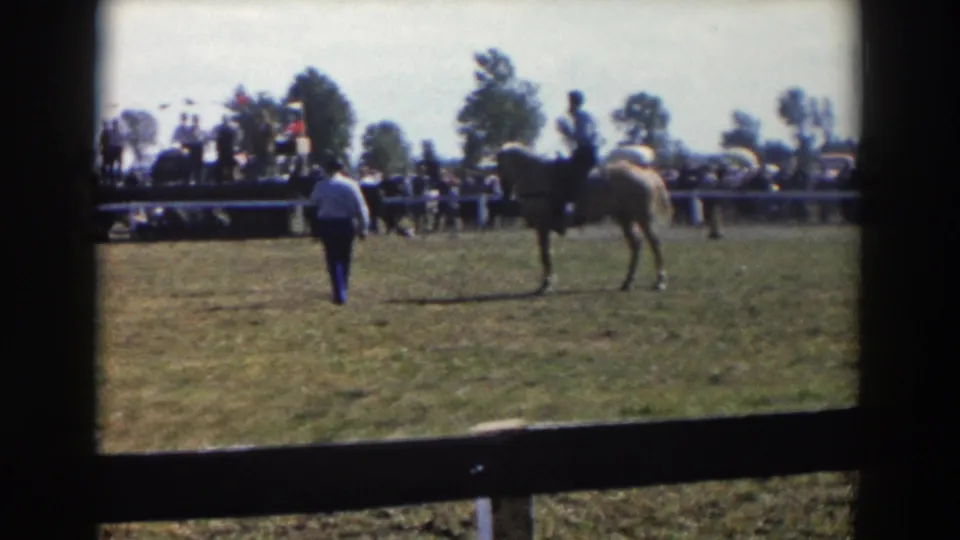A person on foot approaches the field area while a mounted rider remains near the center, with spectators and rail fencing behind.