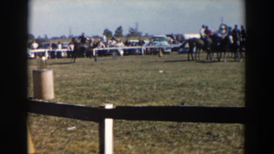 A wide 1957 Wheeling, Ohio home-movie frame shows riders and horses spread across the enclosure with cars and spectators along the edge.