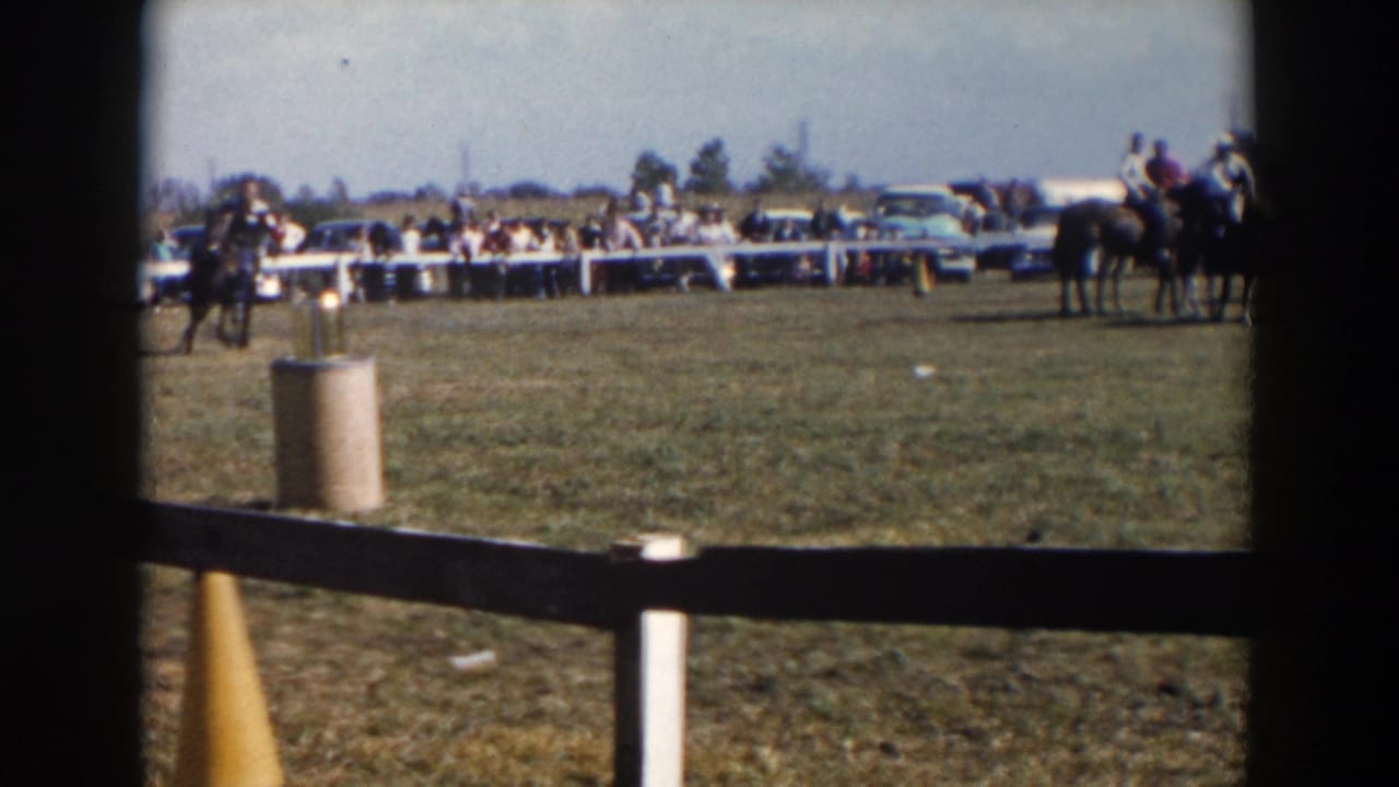 Rider on a horse crosses a grassy rodeo field with spectators and parked cars behind a fence.