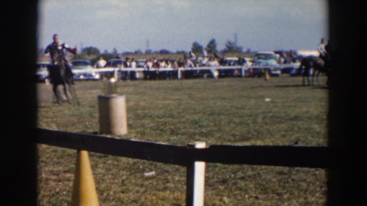 Cowboy rider approaches a course marker on a grassy field with cars and spectators beyond the fence.