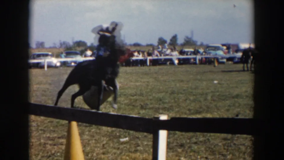 Close pass of a dark horse and rider in the 1957 Wheeling, Ohio rodeo footage, seen from behind a low rail with cars and spectators beyond.