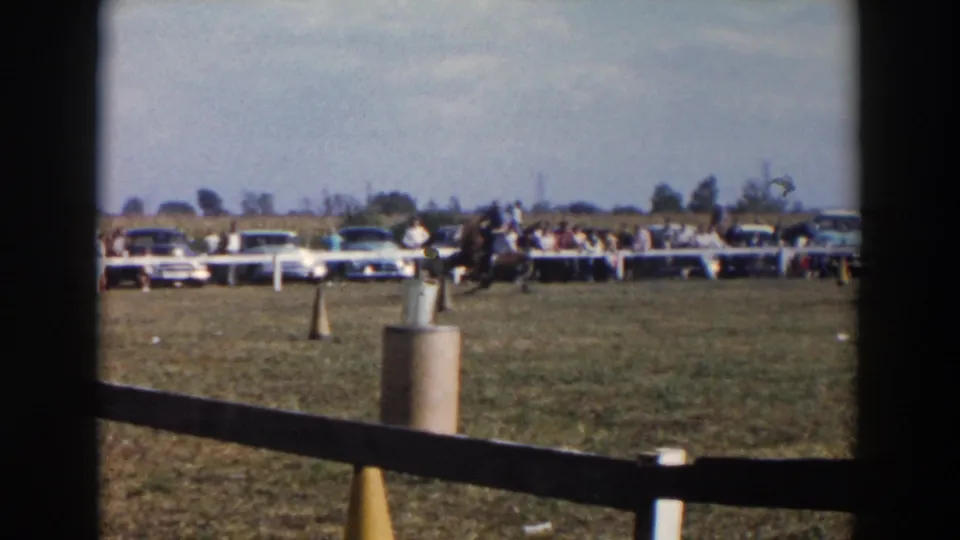 Wide 1957 Wheeling, Ohio rodeo field view with a foreground rail, course markers, spectators behind a white fence, and parked mid-century cars.