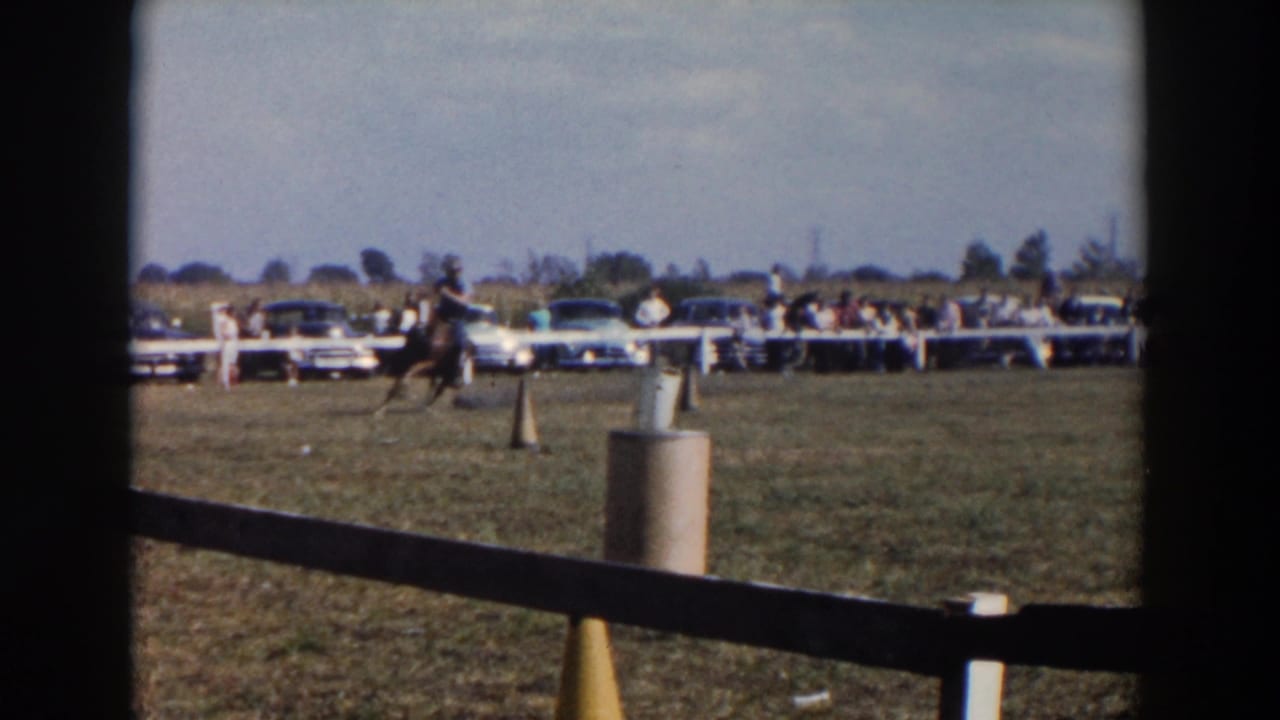 Cowboy on horseback rides across a grassy field near cones, fencing, spectators, and parked cars.