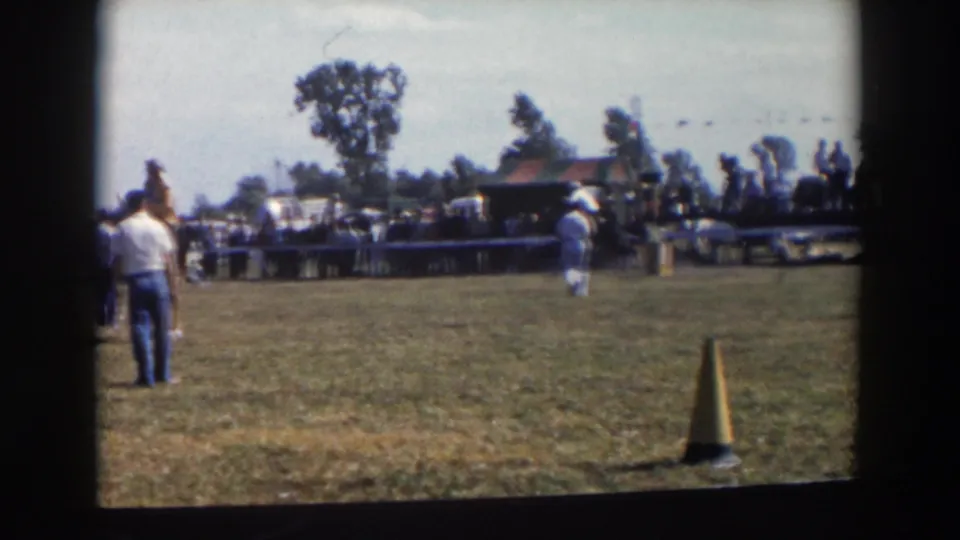A white-shirted spectator or handler stands at the edge of the 1957 Wheeling, Ohio riding field while a rider passes near a yellow cone.
