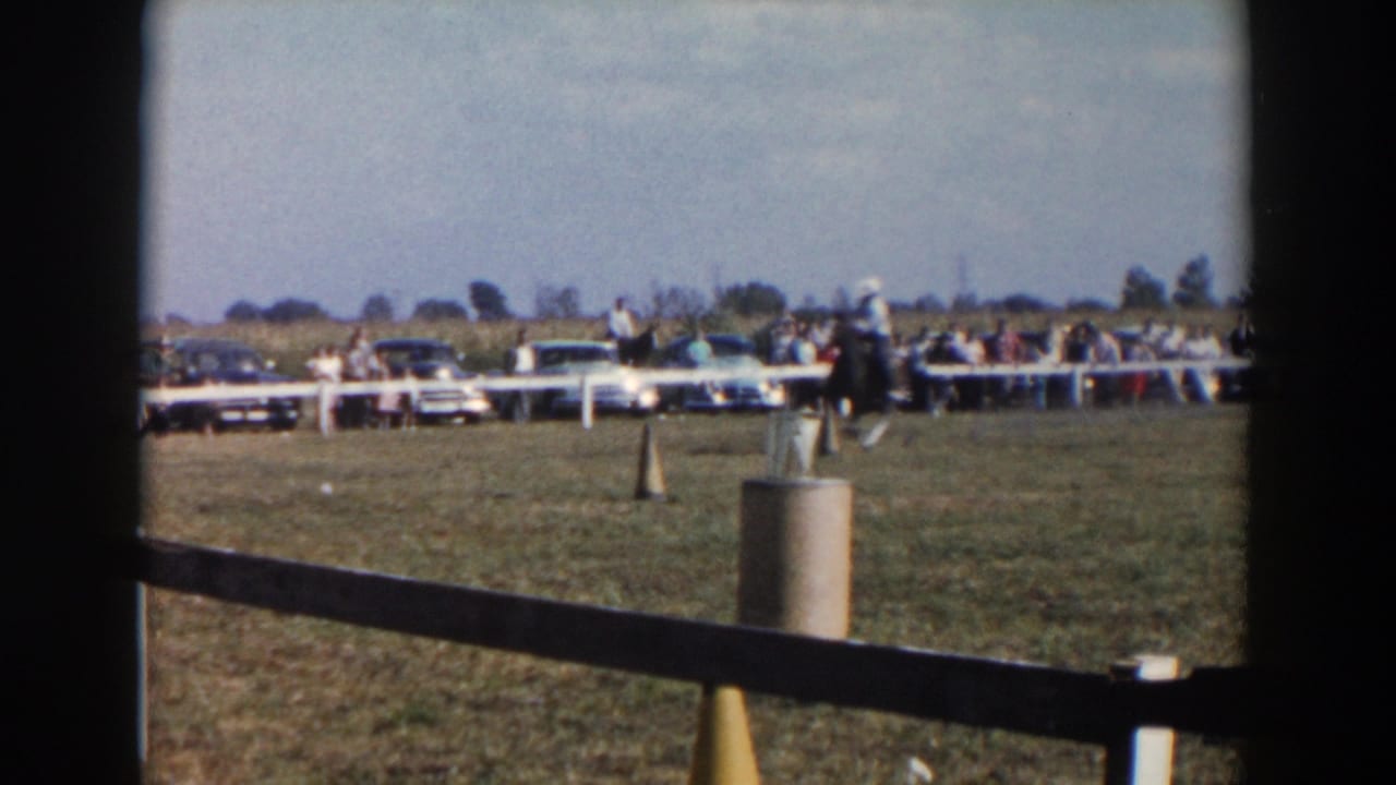 In the supplied 1957 Wheeling, Ohio context, a rider on a dark horse approaches course markers while spectators and parked cars line the field.