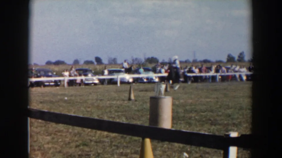 In the supplied 1957 Wheeling, Ohio context, a rider on a dark horse approaches course markers while spectators and parked cars line the field.