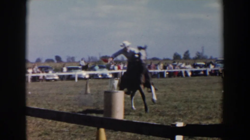 The rider and dark horse pass close to a barrel or marker, with 1950s spectators and cars visible behind the white barrier.