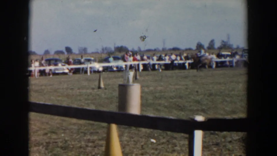 The horse and rider continue across the Wheeling, Ohio course, with cones, a foreground fence, spectators, and mid-century cars in view.