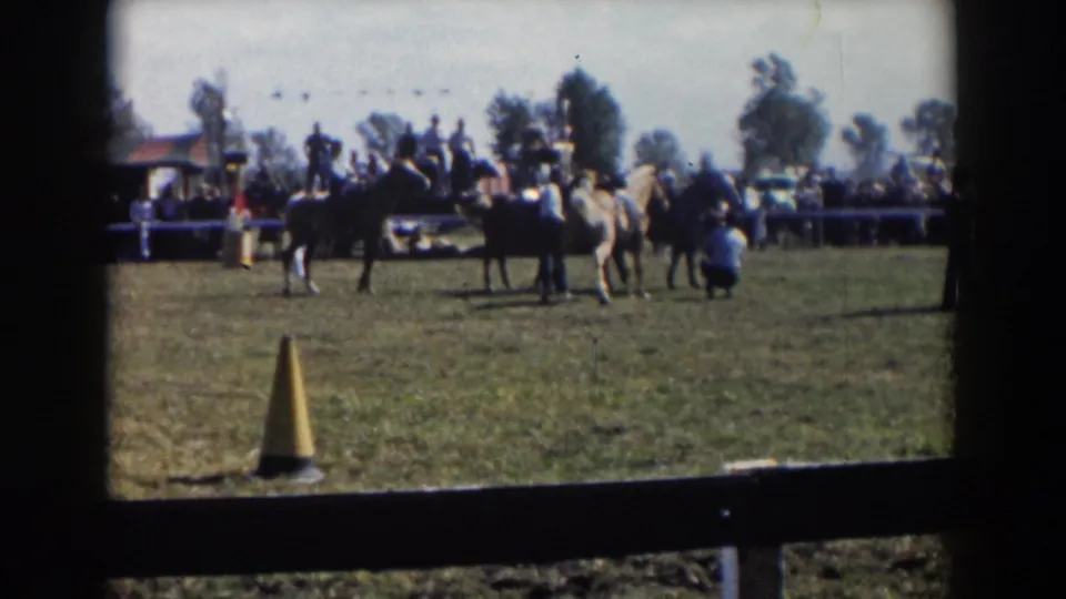 1957 Wheeling, Ohio 8mm frame showing riders on horses in a grass field with spectators, bunting, trees, and a yellow cone.