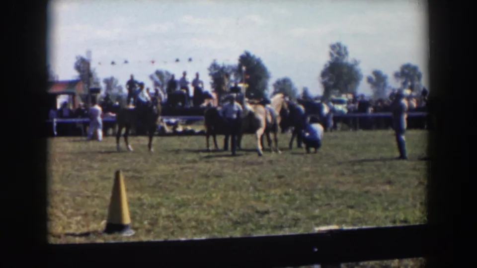 1957 Wheeling, Ohio 8mm frame showing riders with horses, a crouching handler or spectator, a yellow cone, fence-line spectators, and trees.