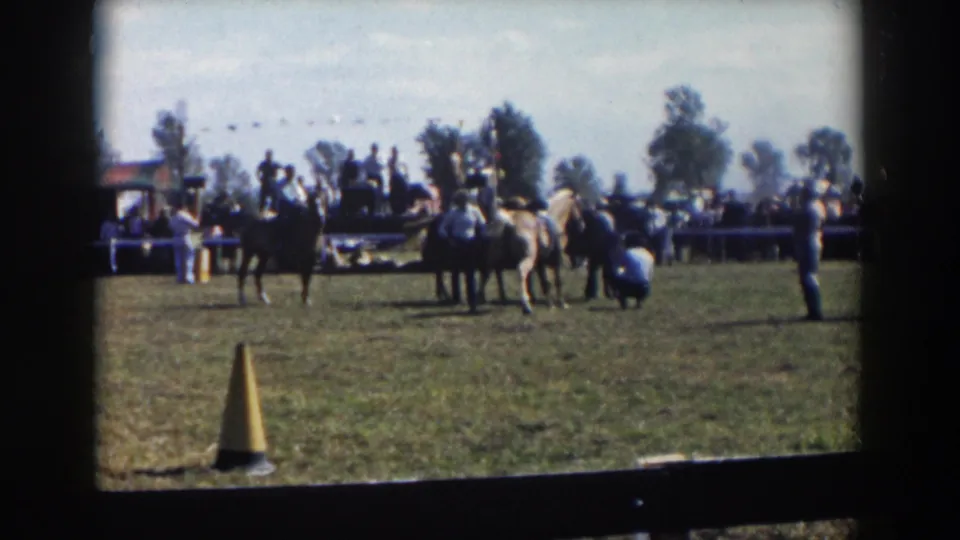 1957 Wheeling, Ohio frame of riders and horses on grass, spectators along the fence, a yellow cone in the foreground, and dark 8mm frame edges.