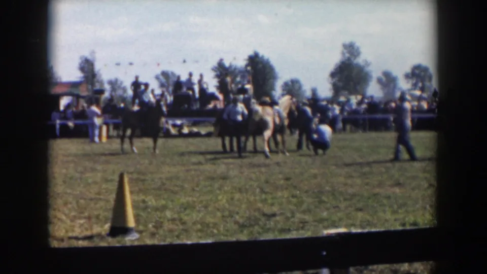 1957 Wheeling, Ohio home-movie frame showing horseback riders, spectators, bunting, parked cars, and a yellow cone on a grassy field.