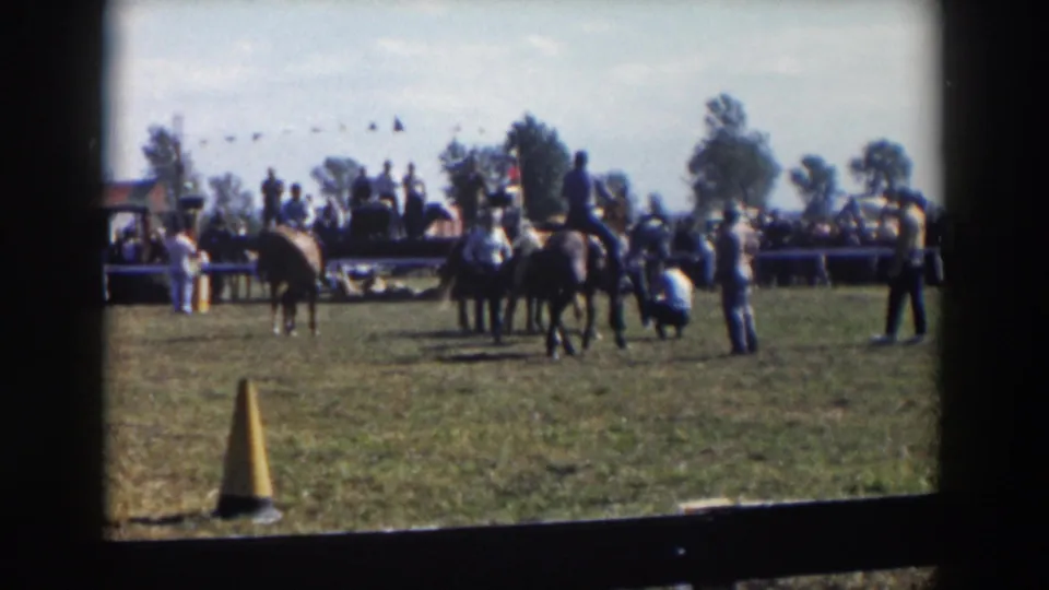 1957 Wheeling, Ohio frame showing riders and horses paused near spectators, with a yellow cone, bunting, cars, and trees behind them.
