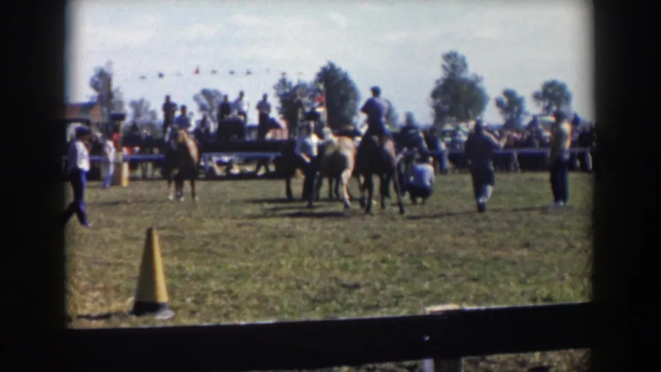 Wide 1957 home-movie view of a Wheeling, Ohio rodeo area with horses, riders, spectators, fencing, and parked cars.