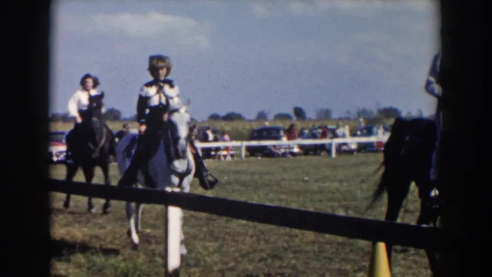 1957 Wheeling, Ohio home-movie frame showing riders on horses near a low fence with spectators and cars beyond the arena.