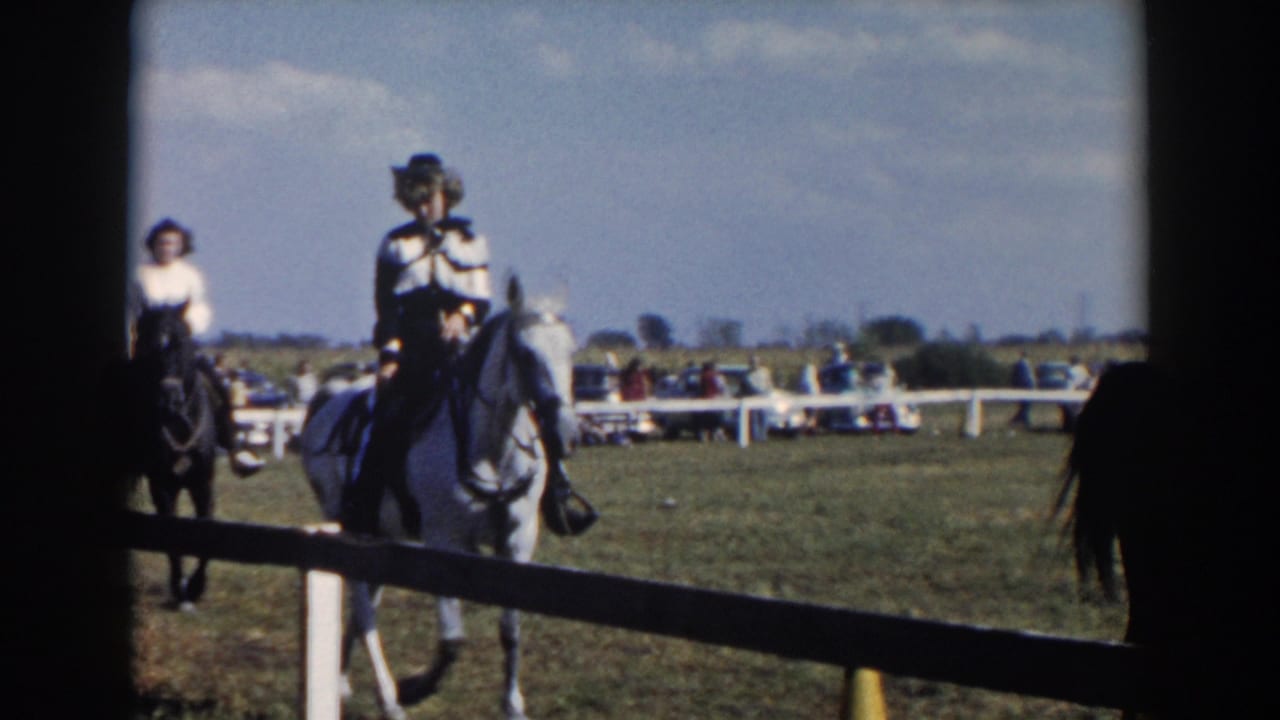 Two riders on horses move along a grassy show ring with spectators and cars behind a white fence.