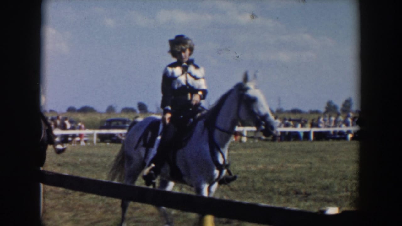 A rider on a white horse passes near the rail while spectators and cars sit beyond the ring.
