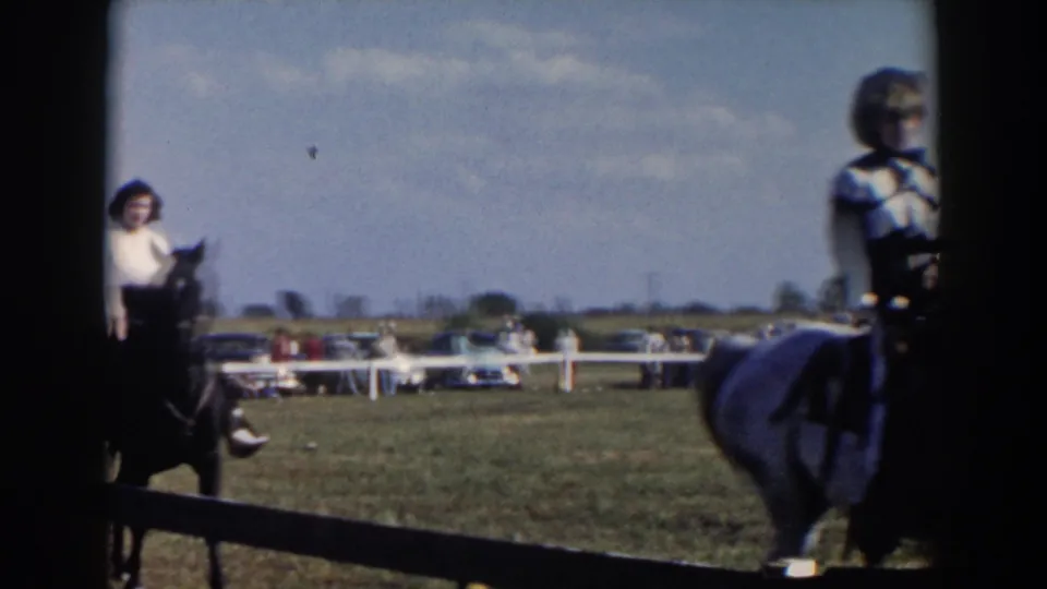 Home-movie frame showing riders crossing the camera's view in a grassy Wheeling, Ohio equestrian ring.
