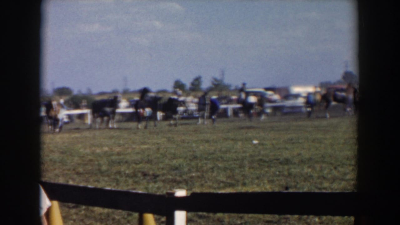 Horses and riders cross a grassy enclosure behind a dark foreground fence.