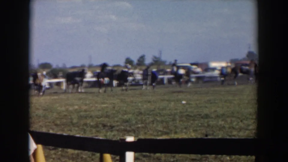 1957 Wheeling, Ohio home-movie frame showing horses with riders in a grassy enclosure, with spectators and cars in the distance.