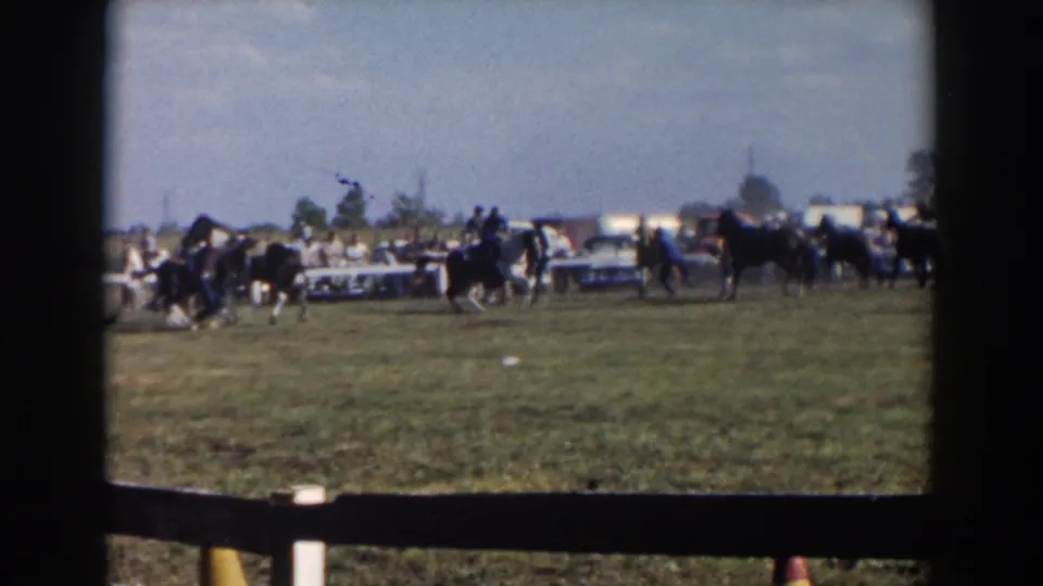 A 1957 8mm frame shows riders trotting horses across a grassy Wheeling enclosure, filmed from a spectator position behind fencing.