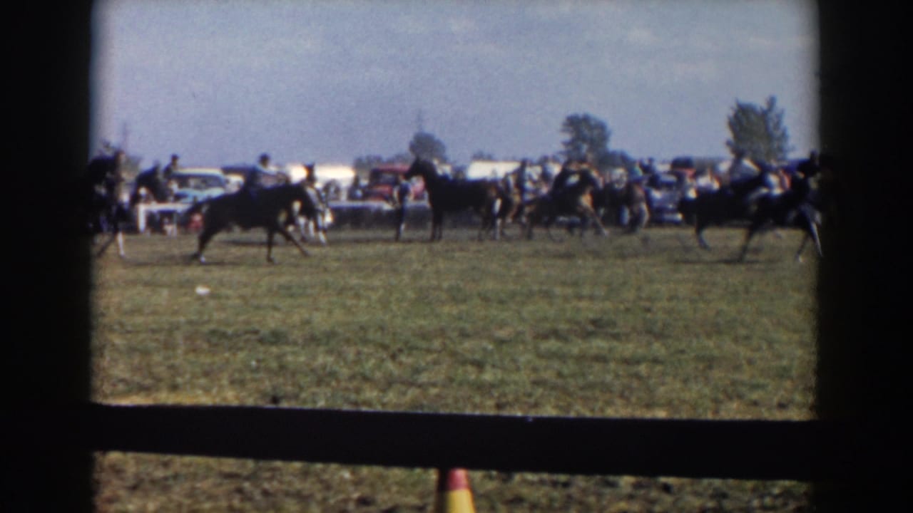 Riders on horses cross a grassy field as spectators and parked cars line the background.