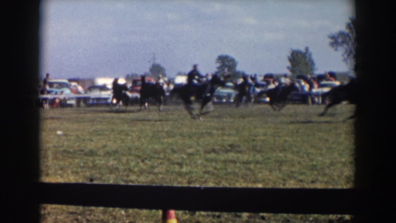 Horseback riders move across the field with a line of spectators and cars behind a barrier.