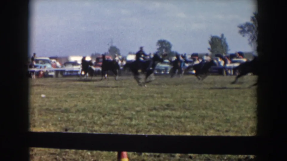 Horseback riders gallop through a grassy Wheeling, Ohio field in 1957, with spectators and parked automobiles visible in the background.