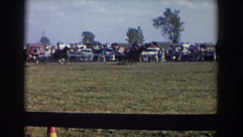 A rider on a dark horse crosses the open field during a 1957 Wheeling, Ohio riding event, with spectators and cars beyond the rail.